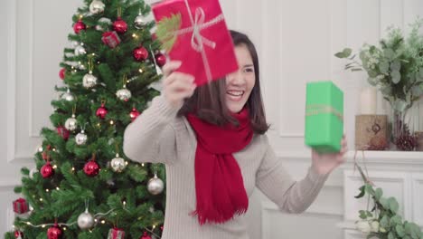 Cheerful-happy-young-Asian-woman-holding-christmas-gifts-smiling-to-camera-in-her-living-room-at-home-in-Christmas-Festival.-Lifestyle-woman-celebrate-Christmas-and-New-year-concept.