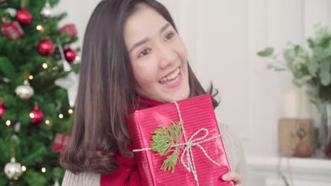 Cheerful-happy-young-Asian-woman-holding-christmas-gifts-smiling-to-camera-in-her-living-room-at-home-in-Christmas-Festival.-Lifestyle-woman-celebrate-Christmas-and-New-year-concept.