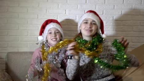 Happy-family,-two-sisters,-in-Santa's-hats-playing-with-colorful-tinsel,-sit-and-dance-on-the-couch-against-a-white-brick-wall,-look-at-the-camera-and-smile.