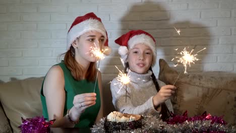 Happy-children-in-Santa's-hats-with-lighted-sparklers-play-sitting-on-the-couch-near-the-cake-and-smile.