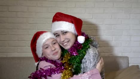 Two-cute-young-girls-in-Santa-Claus-hats-and-Christmas-tinsel-on-their-shoulders-hugging-sitting-on-the-couch,-laughing-and-looking-at-the-camera.-Portrait.-Close-up.-4K.-25-fps.