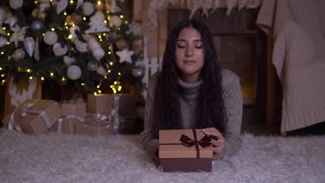 The-girl-opens-the-box-with-a-gift-and-rejoices-lying-on-the-floor-near-the-Christmas-tree.-4K