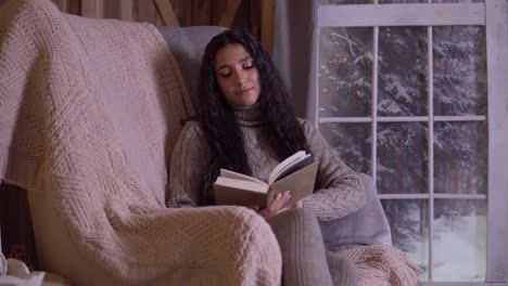 A-young-girl-is-reading-a-book-while-sitting-in-a-chair-near-the-Christmas-tree.