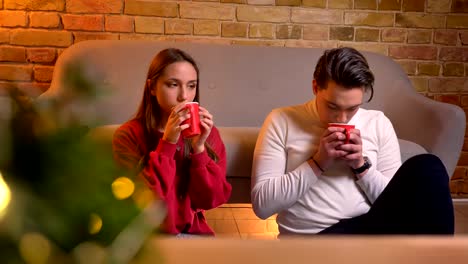 Close-up-portrait-of-talking-young-caucasian-friends-sitting-on-carpet-and-drinking-hot-beverages-in-christmas-atmosphere.