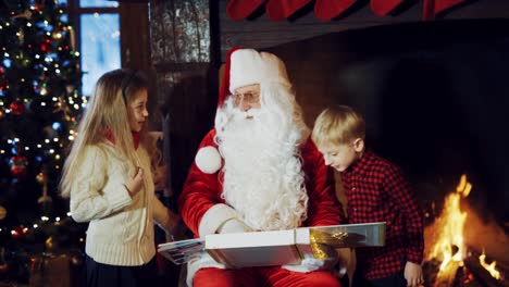 Santa-Claus-is-sitting-in-the-middle-of-a-room-with-an-album-in-his-hands,-surrounded-by-children-on-the-background-of-a-fireplace,-and-telling-a-New-Year-story.