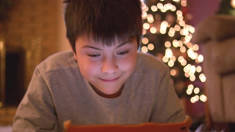 Niño-tendido-en-el-suelo-usando-su-tableta,-sonríe-divertido-a-la-cámara,-árbol-de-Navidad-con-luces-detrás-de-él