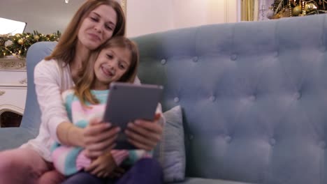 Mutter-mit-Tochter-auf-Couch-mit-Tablet-Computer-Happy-Smiling-junge-Familie-in-der-Nähe-dekorierten-neuen-Jahr-Weihnachtsbaum