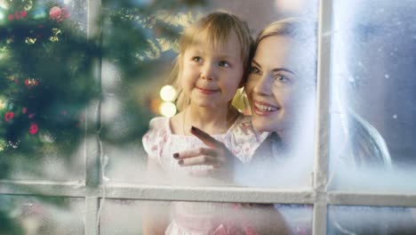 An-Heiligabend-suchen-Mutter-und-Tochter-durchs-Schneefenster-Garland-leuchtet-auf-einem-Fenster