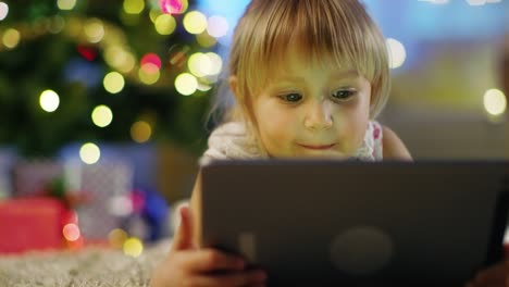 Cute-Little-Girl-With-Tablet-Computer-in-Her-Hands-Lies-on-the-Carpet-under-the-Christmas-Tree.