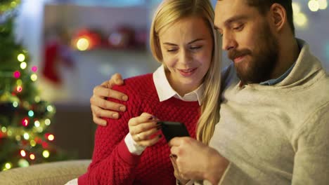 Gut-aussehender-Mann-und-schöne-Frau-sitzen-auf-der-Couch-mit-Smartphone.-Mann-hält-seine-Dame.-In-den-Hintergrund-viele-Lichter-und-Weihnachtsbaum.