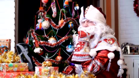Santa-Claus,-Weihnachtsmann,-Väterchen-Frost-arbeitet-an-einem-Laptop-an-einem-Tisch,-unter-Weihnachten-präsentiert,-im-Hintergrund-ist-Weihnachtsbaum