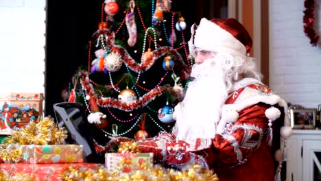 Santa-claus,-father-christmas,-father-frost-is-working-on-a-laptop-at-a-table,-among-christmas-presents,-in-the-background-is-christmas-tree