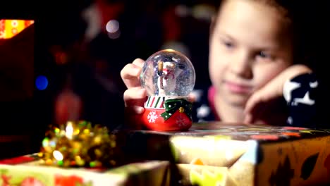 In-the-twilight-of-the-night,-among-the-gifts-in-bright-colorful-paper-packages,-a-pretty-little-blonde-girl-with-a-pink-bow-in-her-hair-and-a-beautiful-dress,-looks-carefully-and-admires-snow-globe.-toy-Christmas-ball-with-sparkles-and-a-snowman-inside