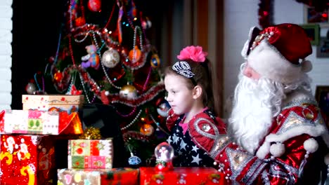 In-der-Dämmerung-der-Nacht-unter-die-Geschenke-in-leuchtend-bunten-Papier-Pakete,-die-hübsche-blonde-Mädchen-mit-rosa-Schleife-im-Haar-und-in-einem-schönen-Kleid-und-Santa-Claus,-Weihnachtsmann,-Väterchen-Frost-beobachten-etwas-fröhlich-auf-einem-Laptop.-Sie-sprechen