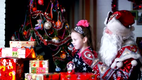 In-the-twilight-of-the-night,-among-the-gifts-in-bright-colorful-paper-packages,-the-pretty-blonde-girl-with-a-pink-bow-in-her-hair-and-in-a-beautiful-dress-and-Santa-Claus,-Father-Christmas,-Father-Frost-watching-something-cheerful-on-a-laptop.-They-talk