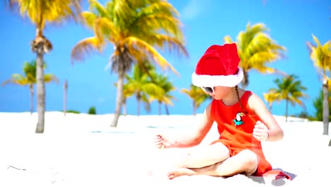 Kleine-entzückende-Mädchen-in-roten-Santa-Hut-am-tropischen-Strand-an-Denweihnachtsfeiertagen