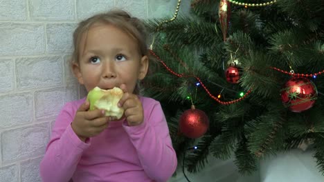 Niedliche-kleine-Mädchen-sitzen-auf-dem-Boden-und-Essen-Apfel-neben-einem-Weihnachtsbaum.