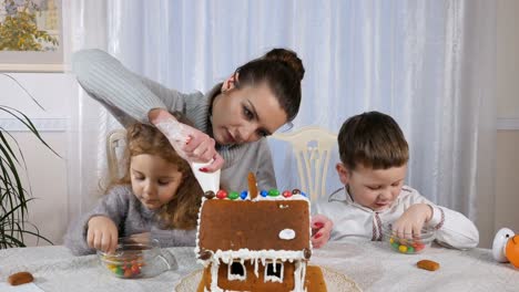 Junge-Mutter-mit-kleinen-Kindern-schmückt-ein-Lebkuchenhaus-mit-einer-Creme