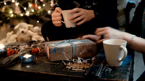 Vista-de-cerca-de-pareja-de-jóvenes-sentados-en-la-mesa,-cerca-del-árbol-de-Navidad-y-abrir-las-fiestas-presentes,-tomando-té