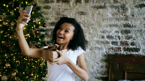 Chica-divertida-carrera-mixta-selfie-fotografiando-en-la-cámara-del-smartphone-en-el-país-cerca-de-árbol-de-Navidad