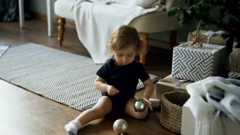 Entzückende-kleine-Mädchen-spielen-mit-Spielzeug-Kugeln-sitzen-in-der-Nähe-von-Weihnachtsbaum-zu-Hause