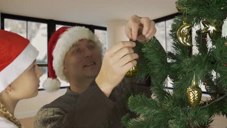 Young-caucasian-girl-daughter-with-father-in-red-Santa-hat-decorate-green-christmas-tree-gold-ball.-Prepairing-Merry-Christmas.-Living-room-window-snowfall.-Rural-countryside.-Snowing-winter-weather.