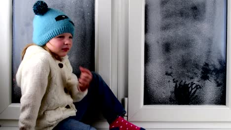 Kleine-Mädchen-sitzt-auf-der-Fensterbank-hinter-dem-gefrorenen-Fenster.-Niedrige-DOF.