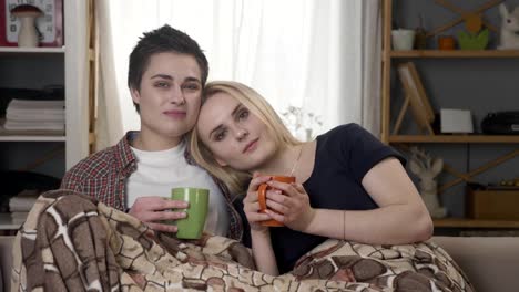 Two-young-lesbian-girls-are-sitting-on-the-couch,-covered-with-a-warm-blanket,-holding-cups-in-their-hands,-cuddling,-looking-at-the-camera-60-fps