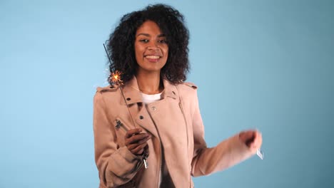 Sparkling-Bengal-fire-in-african-american-woman's-hand-on-blue-background.-Christmas-Holiday-Concept.-Young-pretty-girl-with-afro-hairstyle-celebrating,-smiling,-enjoying-time.-4k