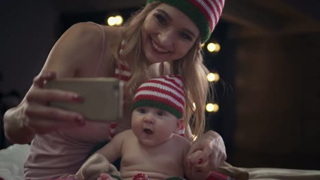 Happy-mother-and-son-in-striped-holiday-caps-are-sitting-on-bed,-using-app-on-smartphone,-smiling-and-laughing