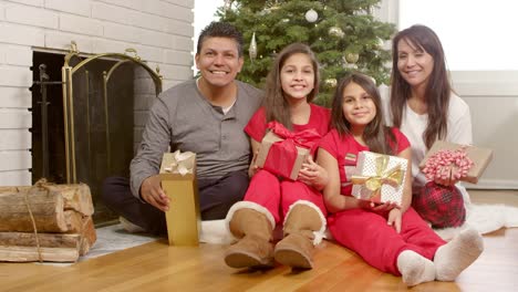 A-family-sits-under-their-christmas-tree-holding-their-presents-and-smiling-into-the-camera