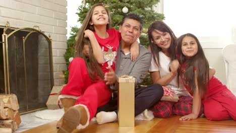 A-man-and-woman-sit-in-front-of-a-christmas-tree-with-presents-and-are-then-are-joined-by-their-two-daughters