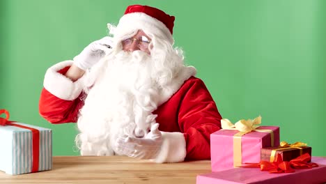 Satisfecho-Papá-Noel-habla-por-teléfono,-se-ríe,-se-sienta-en-la-mesa-con-regalos,-verde-chromakey-en-el-fondo