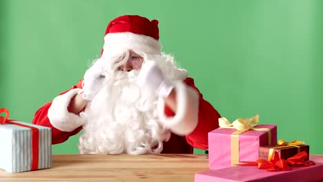 Böser-Weihnachtsmann-am-Telefon-spricht,-schlägt-seine-Faust-auf-den-Tisch,-sitzt-am-Tisch-mit-Geschenken,-grüne-Chromakey-im-Hintergrund