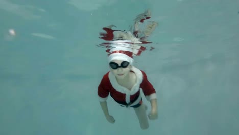 A-happy-little-boy-in-a-suit-and-a-red-Santa-Claus-hat-is-submerged-in-the-pool.-The-boy-is-swimming,-smiling-and-looking-into-the-camera.