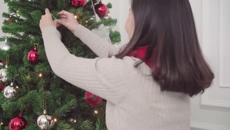 Cheerful-happy-young-Asian-woman-decorating-christmas-tree-in-her-living-room-at-home-in-Christmas-Festival.-Lifestyle-women-celebrate-Christmas-and-New-year-concept.