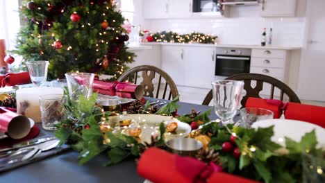 Eine-dekorierte-Weihnachtstisch-mit-Christmas-Cracker-auf-Platten-in-einem-Speisesaal-angeordnet,-ein-Weihnachtsbaum-mit-Küche-im-Hintergrund