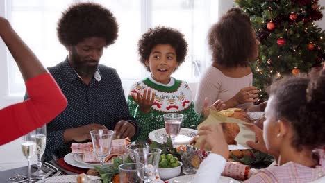 Multi-generation-mixed-race-family-sitting-at-Christmas-dinner-table-wearing-paper-crowns,-panning-shot