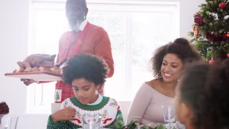 Grandfather-carrying-a-roast-turkey-to-the-dinner-table-during-a-multi-generation,-mixed-race-family-Christmas-celebration,-handheld-tracking-shot