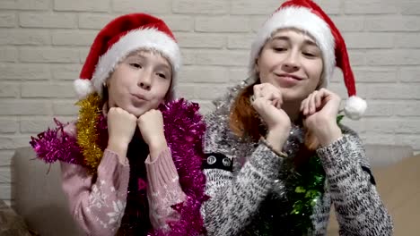 Two-happy-girls-in-Santa-hats-and-tinsel-on-their-shoulders-smile-and-dance,-sitting-at-a-table-against-a-white-wall.-Portrait.