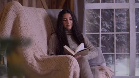 Young-girl-with-long-hair-reads-a-book-while-sitting-in-a-chair-near-the-Christmas-tree.