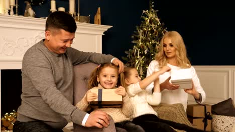 Merry-Christmas-and-Happy-Holidays!-Cheerful-mom-and-dad-and-cute-daughters-girls-exchanging-gifts.-Parent-and-two-little-children-having-fun-and-playing-together-near-Christmas-tree-indoors