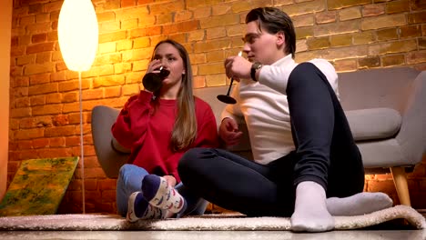 Close-up-portrait-of-young-caucasian-friends-sitting-on-carpet-and-drinking-wine-talking-with-each-other-in-cosy-home-atmosphere.