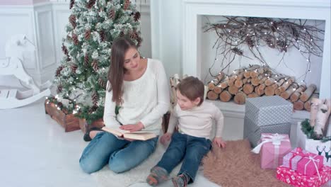 Joven-madre-leyendo-el-libro-para-distraer-a-su-hijo-de-Navidad-presenta