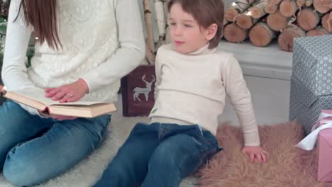 Capricious-boy-listening-to-his-mom-reading-a-book-near-Christmas-tree