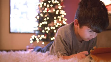 Little-boy-laying-on-the-floor-using-his-tablet,-Christmas-tree-with-lights-behind-him