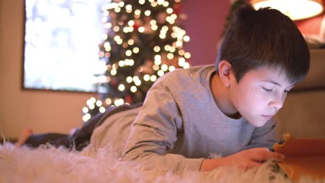 Niño-acostado-en-el-suelo-usando-su-tableta,-árbol-de-Navidad-con-luces-detrás-de-él