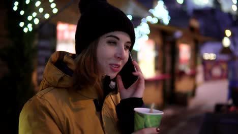 Beautiful-Girl-dialing-a-number,-Talking-with-Friend-and-holding-a-paper-cup-of-hot-tea-at-the-christmas-tree-background-in-the-evening.