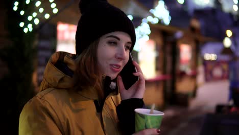 Schöne-Mädchen-wählen-einer-Nummer,-Talking-mit-Freund-und-hält-eine-Papier-Tasse-heißen-Tee-am-Weihnachtsbaum-am-Abend-Hintergrund.