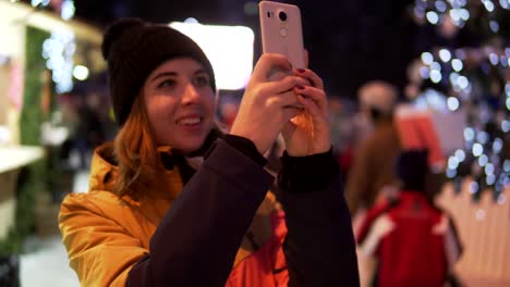 Mujer-joven-en-ropa-de-invierno-brillante-tomar-foto-con-el-teléfono-móvil-en-el-mercado-de-Navidad